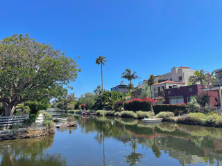 Picturesque view of the Venice Canals featuring calm water, lush greenery, footpaths, and charming colorful houses. Small boats float along the scenic canal under a bright clear sky