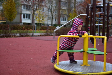 Toddler in a striped snowsuit spinning on a yellow carousel at an outdoor playground during autumn. City park background with trees and soft rubber flooring creates a calm, everyday childhood moment.