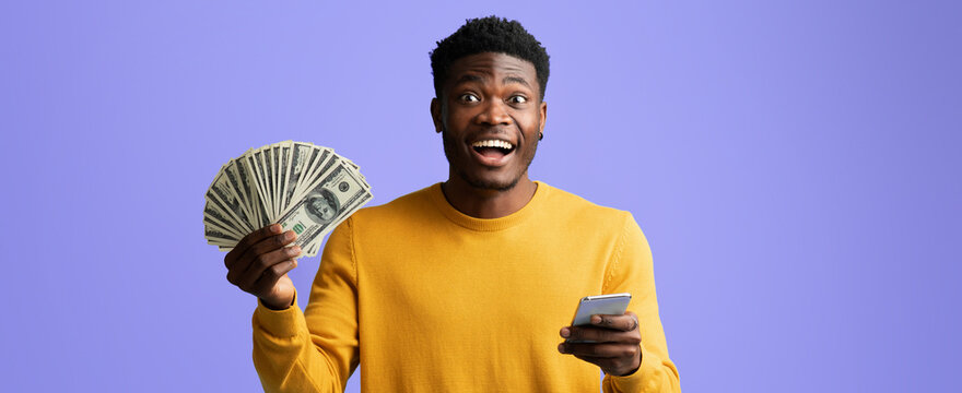A man holds a fan of cash in one hand while using a smartphone in the other. He appears excited and joyful, with a purple background enhancing the festive mood. - Powered by Adobe