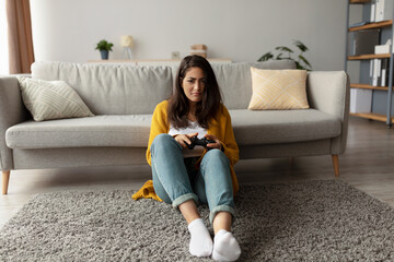 Obraz premium A serious arab woman sits on the carpet in her living room, focused on playing video games with a joystick. The space is neat and inviting, with soft lighting and a sofa in the background.