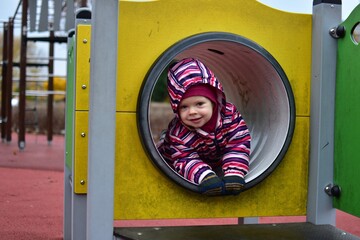 Smiling toddler in a colorful striped snowsuit playing inside a playground tunnel on an autumn day. Bright yellow structure and soft light capture a joyful, candid childhood moment.