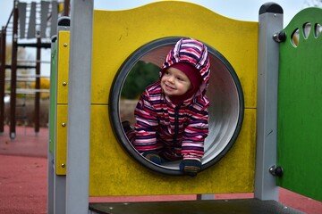 Smiling toddler in a colorful striped snowsuit playing inside a playground tunnel on an autumn day. Bright yellow structure and soft light capture a joyful, candid childhood moment.