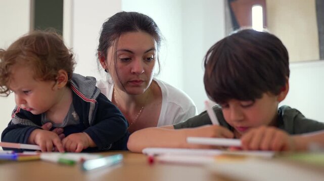 Baby explores markers beside older brother drawing at table while mother remains nearby creating tender family moment showing curiosity concentration and early creative learning in warm home setting