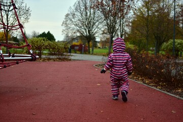 Toddler in a striped snowsuit walking away on a red rubber playground path during autumn. Bare trees, bushes and soft light create a calm, natural outdoor childhood scene.