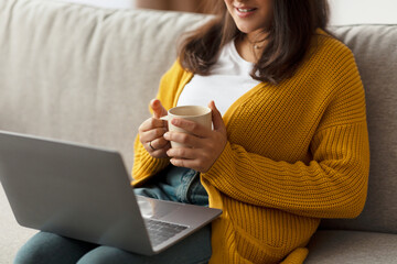 A young Arab woman sits comfortably on her sofa, holding a cup of tea while watching videos on her laptop. She enjoys her leisurely weekend, relaxed and cozy at home.