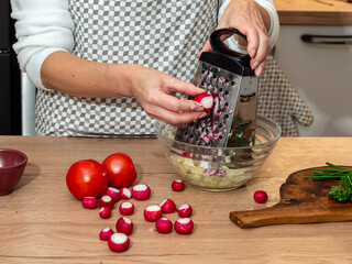 Woman grating fresh radishes while preparing a homemade salad. Vegetables and kitchen utensils lie on the countertop, creating a natural culinary scene.