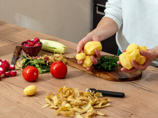 Woman holding freshly peeled potatoes with vegetables and herbs on the kitchen countertop. A natural culinary scene showing home meal preparation.