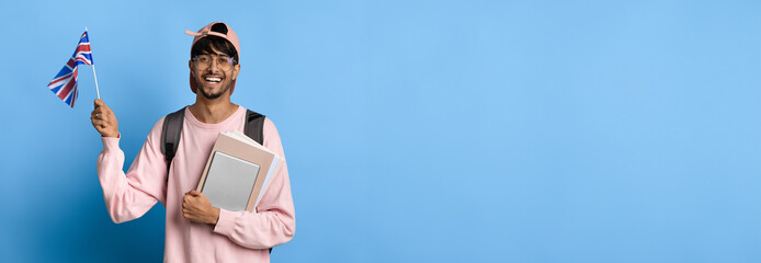 A young man stands against a solid blue background, smiling broadly while holding a small British...