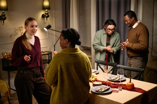 Group of young adult and middle aged multiethnic friends gathering around dining table, talking and laughing during Thanksgiving dinner, standing and interacting before meal