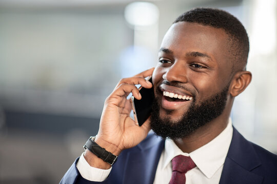 Closeup shot of happy handsome young black businessman in nice suit talking on smartphone with business partner while walking by office building, looking at copy space and smiling, panorama
