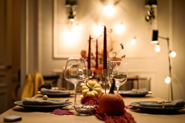 Thanksgiving dinner table setting featuring empty wine glasses, plates, napkins, decorative pumpkins and candles arranged on table, no people visible, warm indoor lighting