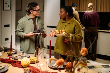 Multiethnic group of young adults standing around table sharing food during Thanksgiving dinner, Caucasian man holding plate of grapes talking to Black woman holding plate of muffins