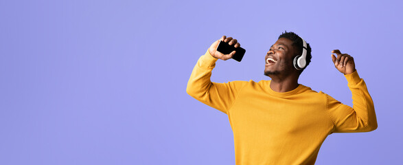 A man is joyfully singing along to music while wearing headphones and holding a smartphone. He appears to be having a great time, dancing against a vibrant purple background.