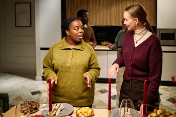 Young adult Black woman and young adult Caucasian woman standing at dining table talking, multiethnic male friends in background preparing food during Thanksgiving dinner among neighbours
