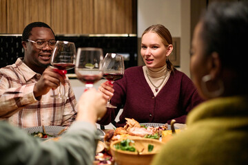 Diverse group of young adults and middle aged woman raising wine glasses during Thanksgiving dinner among friends, smiling and making toast around festive table with food