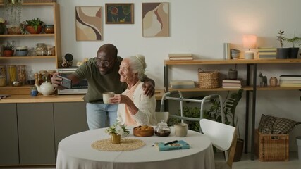 Wide angle shot of senior woman taking selfie photo with Black man as social worker at table in cozy kitchen, copy space - Powered by Adobe