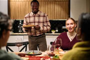 Black middle aged man serving pie to group of diverse young adults and middle aged women sitting around table enjoying thanksgiving dinner together, food and wine visible on table