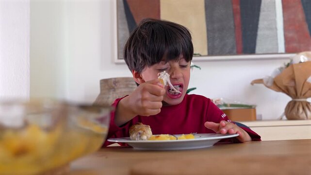 Hungry young boy in red shirt aggressively biting into a chicken drumstick at the family dining table, showing intense expression as he focuses on his food during a casual home mealtime