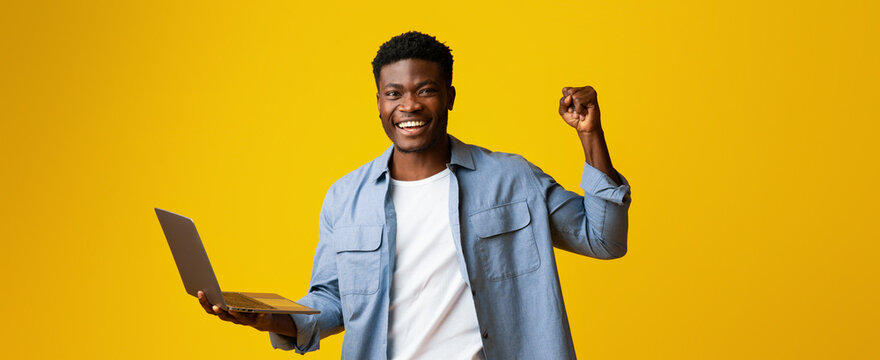 A young man is smiling broadly, raising his fist in joy while holding a laptop in his other hand. He stands against a bright yellow background, exuding happiness and excitement.
