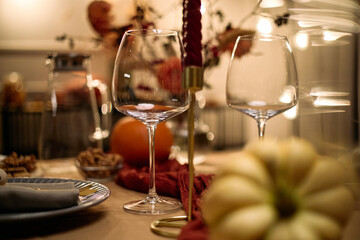Closeup of empty wine glasses standing on festive dinner table with blurred pumpkin, nuts, and autumn floral arrangement in background during Thanksgiving dinner among friends