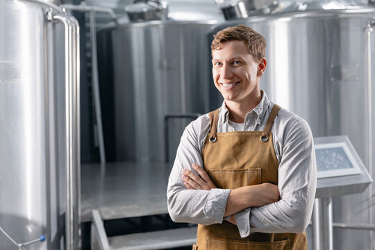 Male brewer in modern brewery wearing apron smiling in front of stainless steel tanks - Powered by Adobe
