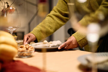 Black woman arranging plates and napkins on dining table during Thanksgiving dinner among friends, hands visible placing tableware, festive meal preparations in progress