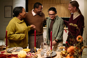 Group of diverse young adults and middle aged woman laughing and talking around festive dinner table during Thanksgiving celebration among friends, smiling and interacting together