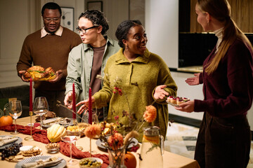 Group of diverse young adults and middle aged woman gathering around table sharing Thanksgiving dinner, smiling and talking, holding plates and serving food, festive meal in modern home