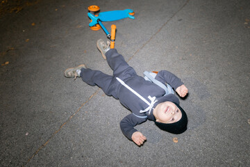 Caucasian male child lying on pavement beside scooter at night, dressed in dark outfit. Lifestyle moment of childhood.