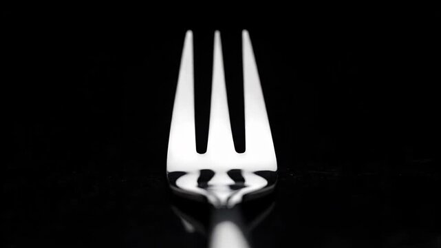 Black and white studio shot of a metal fork with its prongs moving forward on a dark, reflective surface, creating distorted reflections