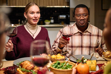 Young Caucasian woman and young Black man sitting at table holding wine glasses, smiling and talking with friends during Thanksgiving dinner, plates of food and drinks visible in foreground