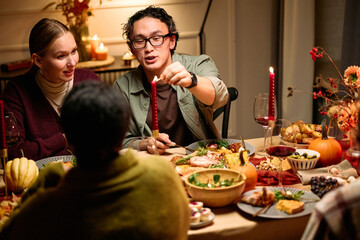 Group of diverse young adults and middle aged woman gathering around table sharing Thanksgiving dinner, smiling and talking, Caucasian man lighting candle, festive meal with traditional dishes