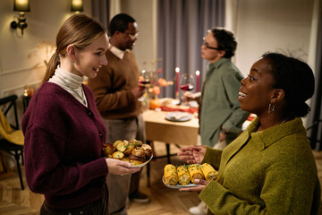 Young adult Caucasian woman and Black woman holding plates of food, smiling and talking, with middle aged Black and Asian males standing near table during Thanksgiving dinner among friends