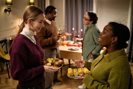 Young adult Caucasian woman and Black woman holding plates of food, smiling and talking, with middle aged Black and Asian males standing near table during Thanksgiving dinner among friends - Powered by Adobe