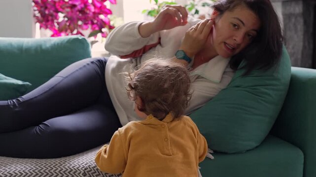 Mother lying on sofa adjusting earring while toddler plays nearby creating natural domestic moment showing care routine and gentle presence shared between parent and young child