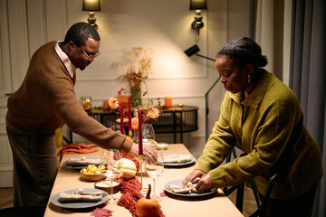 Middle aged Black man and middle aged Black woman arranging plates and napkins on dining table, preparing Thanksgiving dinner among friends, autumn decorations and food visible