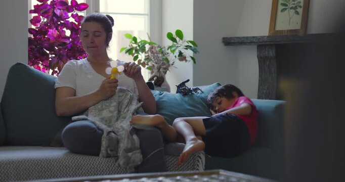 Mother sitting on couch holding breast pump while child rests beside her, creating quiet everyday family moment with calm domestic atmosphere and natural interaction