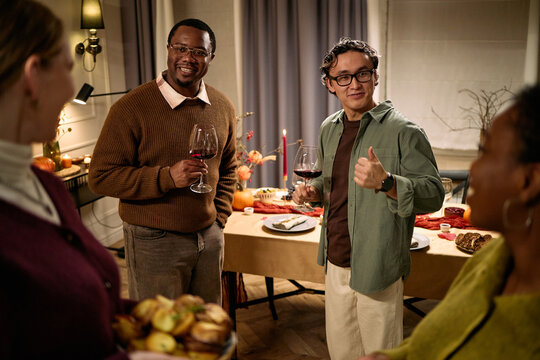 Group of diverse young adults and middle aged Black man standing around table holding wine glasses, smiling and talking during Thanksgiving dinner among friends, food visible in foreground