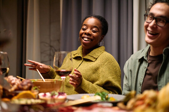 Young Black woman laughing and gesturing while sitting at table with young Caucasian man during thanksgiving dinner among friends, food and wine glasses visible in foreground