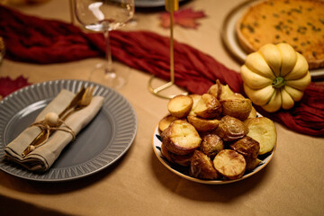 Closeup showing bowl of roasted potatoes on festive table with plate, napkin, cutlery, glass, small pumpkin and pie, capturing detail of Thanksgiving dinner among friends