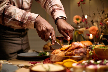 Middle aged Black man carving roasted turkey at dinner table surrounded by traditional Thanksgiving dishes, using knife and fork, celebrating holiday meal with friends