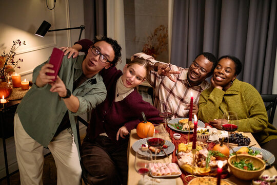 Group of diverse young adults sitting at table taking selfie together during Thanksgiving dinner, smiling and making peace sign gestures, festive food and drinks arranged in front