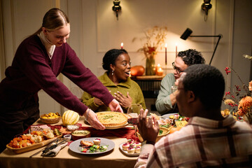 Group of diverse young adults and one Black woman sitting around table sharing Thanksgiving dinner, smiling and talking, Caucasian young woman serving pie to friends, festive food on table
