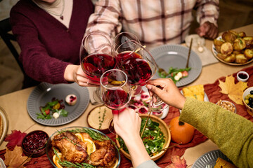 Diverse group of young adult and middle aged friends clinking wine glasses over festive Thanksgiving dinner table, hands of multiethnic men and women holding drinks above holiday meal