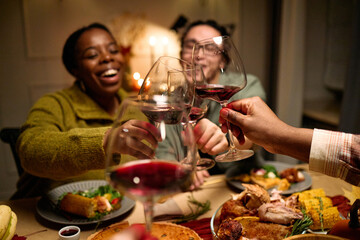 Obraz premium Diverse group of young people and middle aged friends clinking wine glasses while celebrating Thanksgiving dinner, plates with traditional holiday food on table, smiling and enjoying meal together