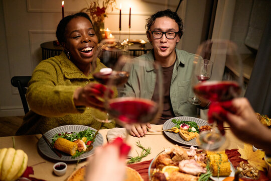 Young adult Black woman and young adult Asian man smiling and raising wine glasses with friends during Thanksgiving dinner, diverse group celebrating around festive table with food