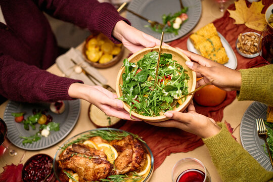 Diverse group of young adult and middle aged hands serving salad at Thanksgiving dinner table, sharing food among friends, visible plates with turkey, vegetables, and festive dishes
