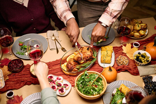 Diverse group of young adult and middle aged friends sharing Thanksgiving dinner, Black man carving roasted turkey while others reaching for food, festive table with salads and wine - Powered by Adobe