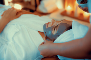 Serene ambiance of spa salon, woman customer indulges in rejuvenating with charcoal face cream massage with warm lighting candle. Facial skin treatment and beauty care concept. Quiescent