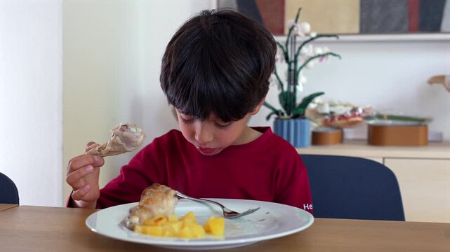 Young boy examining chicken drumstick while eating at home table, looking closely at food with curious expression during a relaxed family mealtime in a warm indoor setting